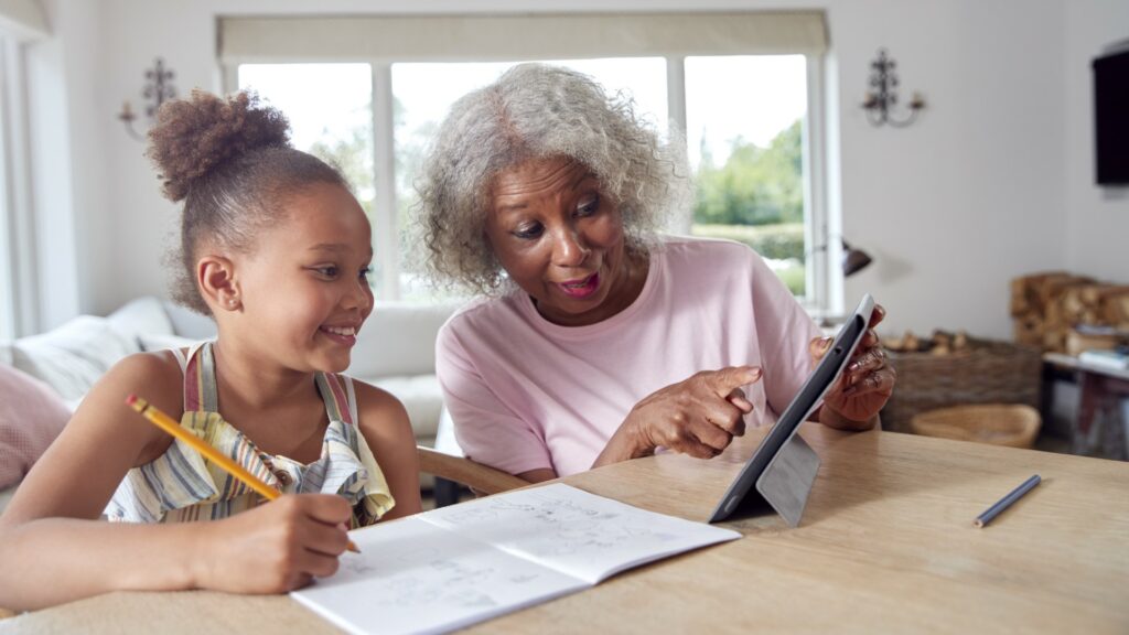 Grandmother Helping Granddaughter With Home Schooling Sitting At Table With Digital Tablet